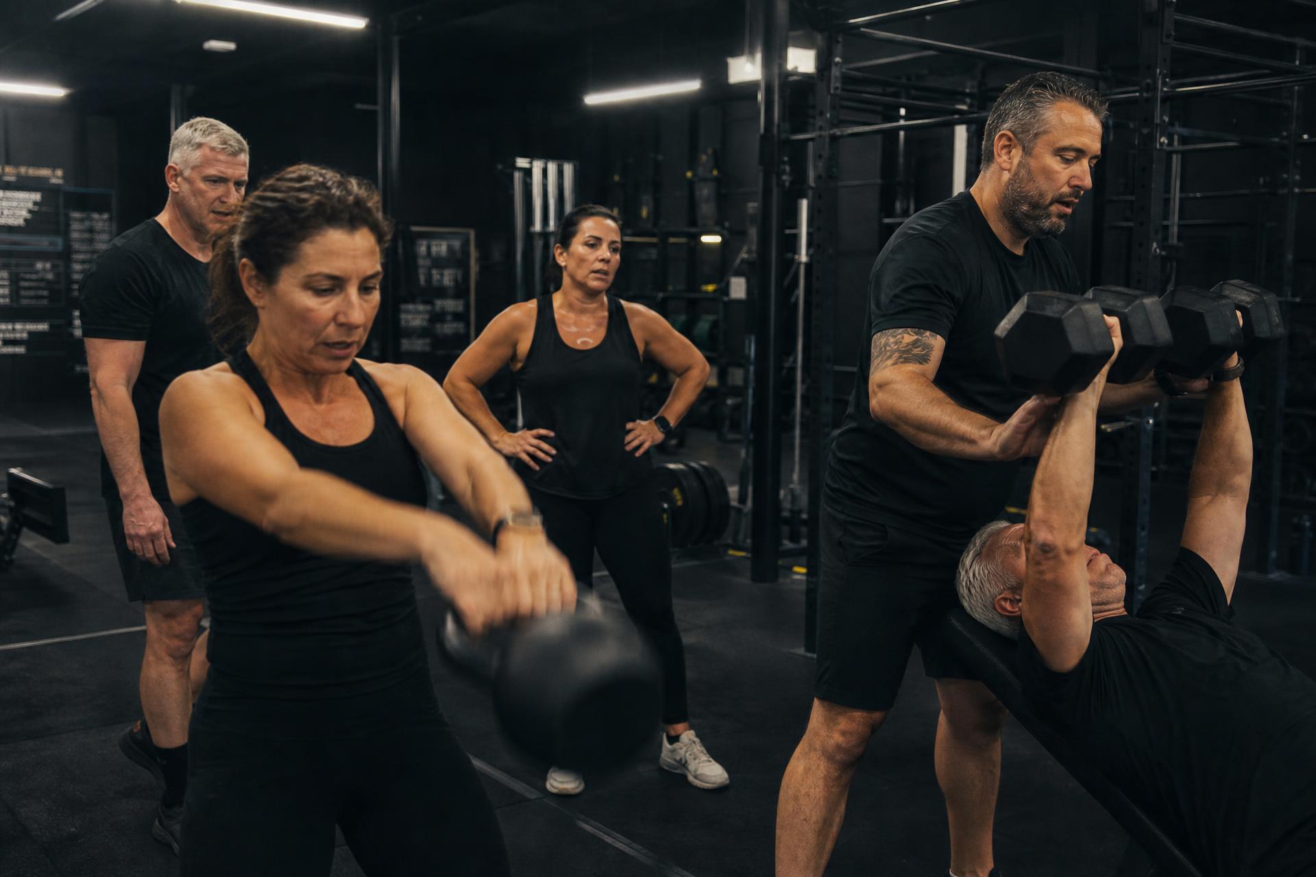 A mixed group of men and women in their 40s and 50s training hard with kettlebells and dumbbells in a real hybrid gym